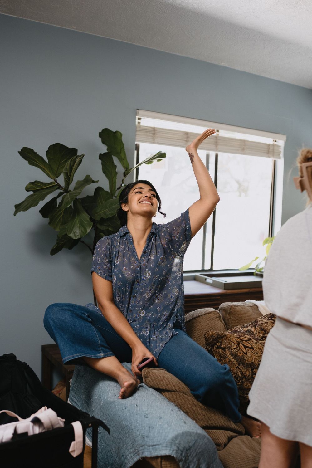 A smiling woman sits cross-legged on a couch, raising her hand while chatting with a friend in a cozy living room.