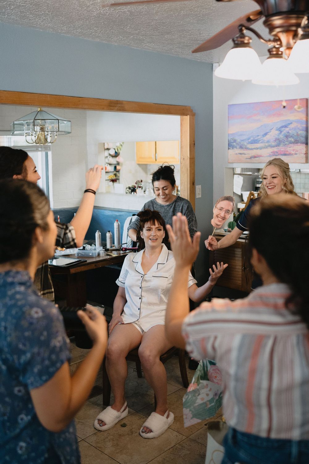 Group of women celebrating together in a home, clapping and smiling around a seated woman in white pajamas.