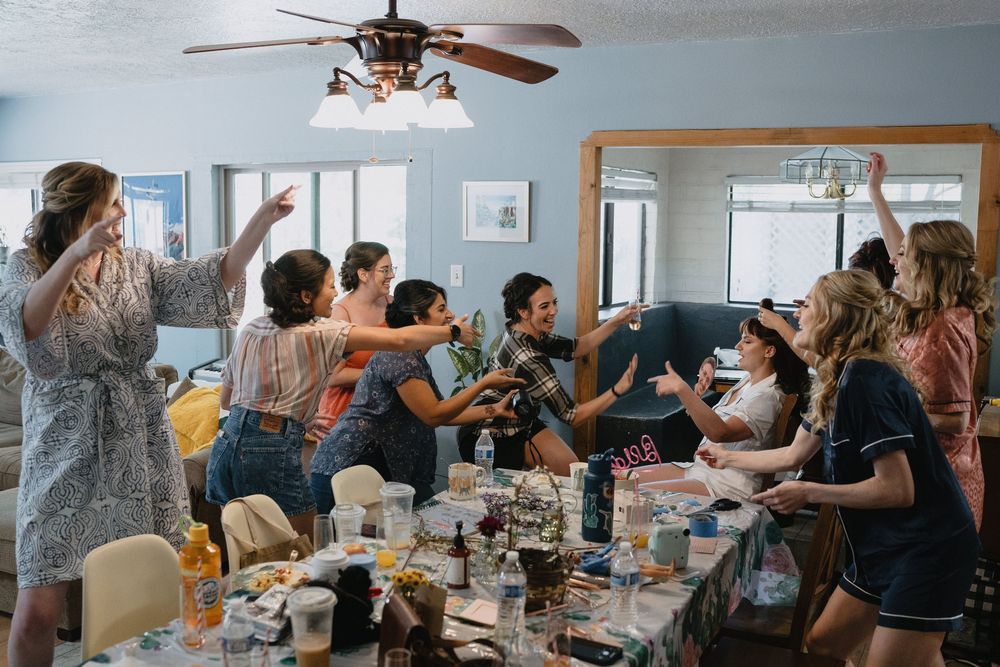 Group of women celebrating at a party, raising hands and dancing around a table filled with food and drinks indoors.