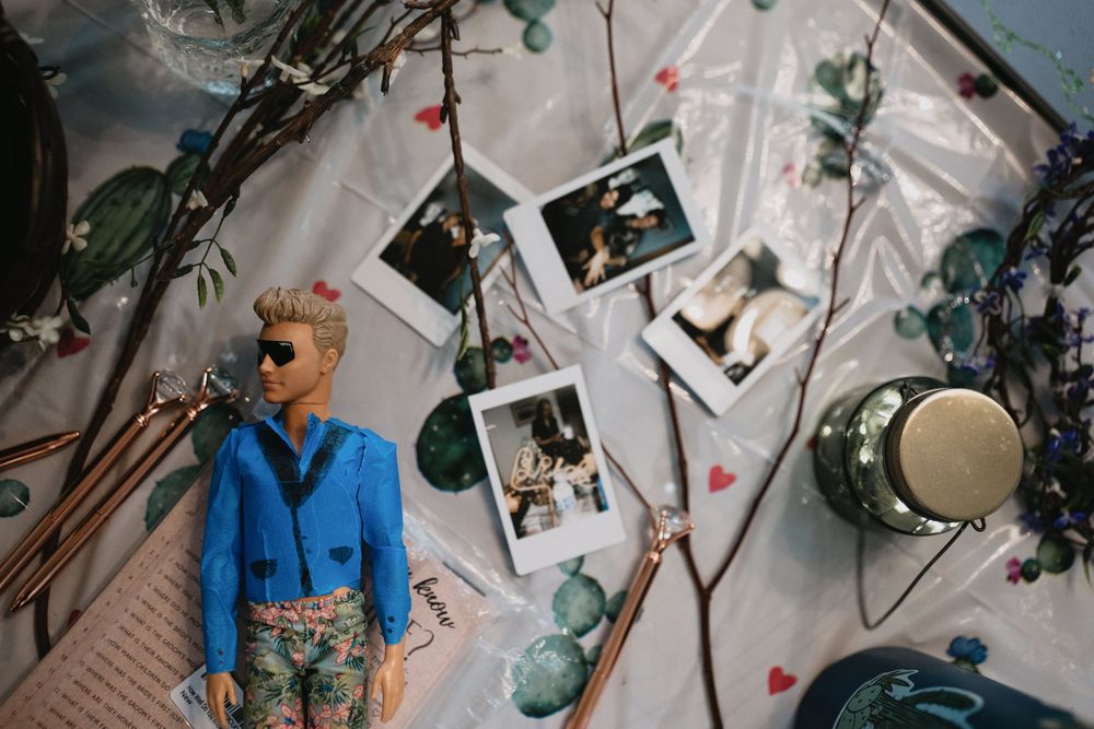 A Ken doll in a blue blazer poses among Polaroid photos, eucalyptus branches, and heart confetti on a white surface.