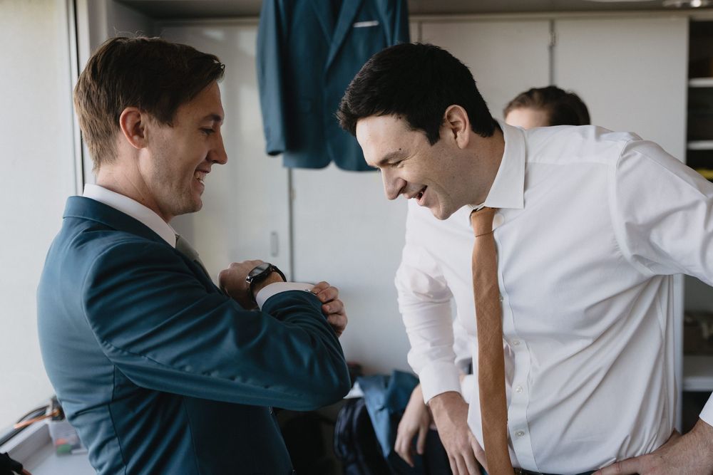 Two groomsmen smiling and helping each other get dressed in suits before a wedding ceremony.