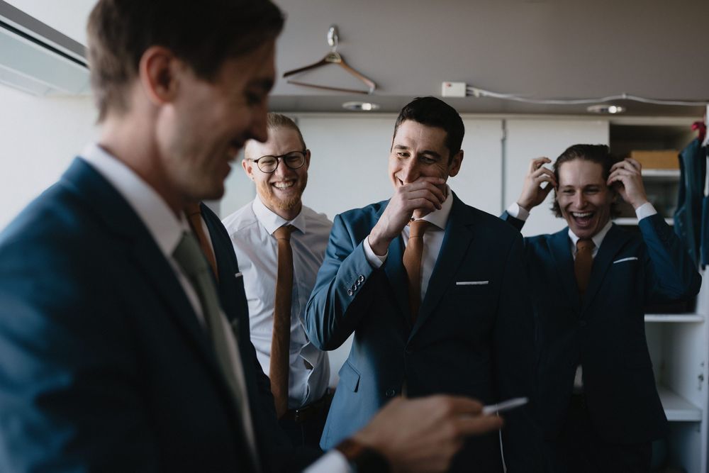 Groomsmen in navy suits laughing and adjusting ties before a wedding ceremony in a bright room.
