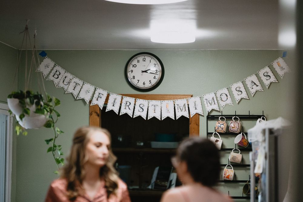 Bridal getting ready room with 'But First Mimosas' banner, wall clock, and blurred bridesmaids in warm lighting.