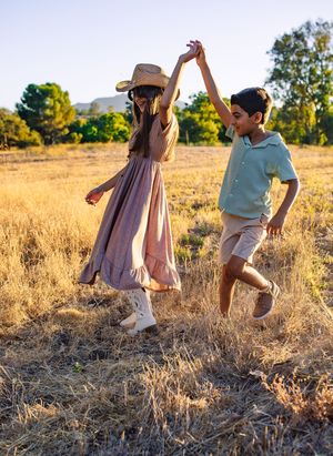 Two children dancing and holding hands in a sunlit golden field, girl in pink dress and cowboy hat.