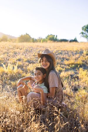 Two smiling children sit together in a golden field at sunset, with the older girl wearing a straw cowboy hat.