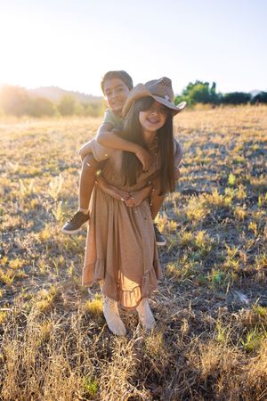 A girl in a cowboy hat gives a boy a piggyback ride in a sunlit golden field at sunset.