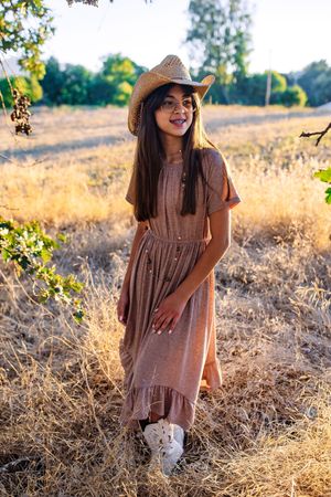 Young girl in brown dress and straw cowboy hat standing in a golden dry field at sunset.