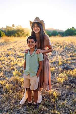 A smiling girl in a cowboy hat and brown dress stands with a boy in a sunny golden field at sunset.