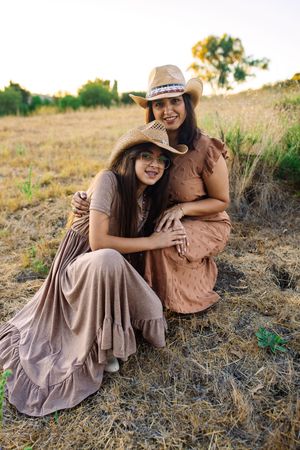 Two women in boho dresses and cowboy hats smile while sitting together in a dry golden field at sunset.