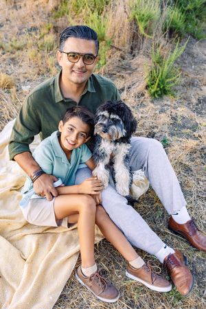 A father and son sit together on a blanket in a dry field, smiling with their fluffy black and white dog.
