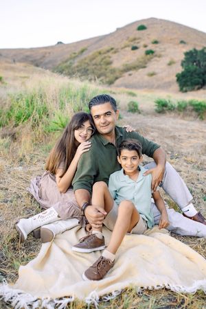 A father sits on a blanket outdoors with his two children, smiling warmly against a scenic hillside backdrop.