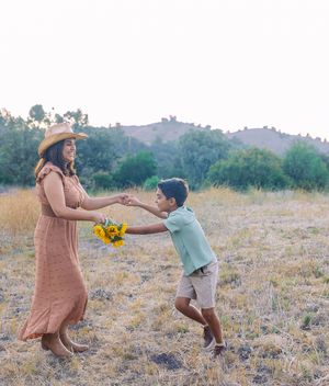 A woman in a straw hat and maxi dress holds sunflowers while dancing with a young boy in a sunny field.