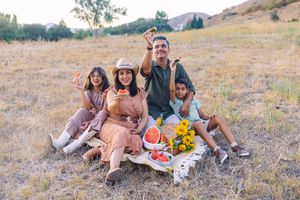 Family of four enjoying a sunny outdoor picnic with watermelon and sunflowers in a golden field.