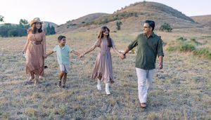 A happy family of four walks hand-in-hand through a scenic grassy hillside on a sunny day.
