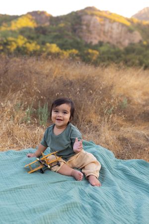 Happy baby sits on a teal blanket in a dry grassy field, playing with a yellow toy airplane outdoors.