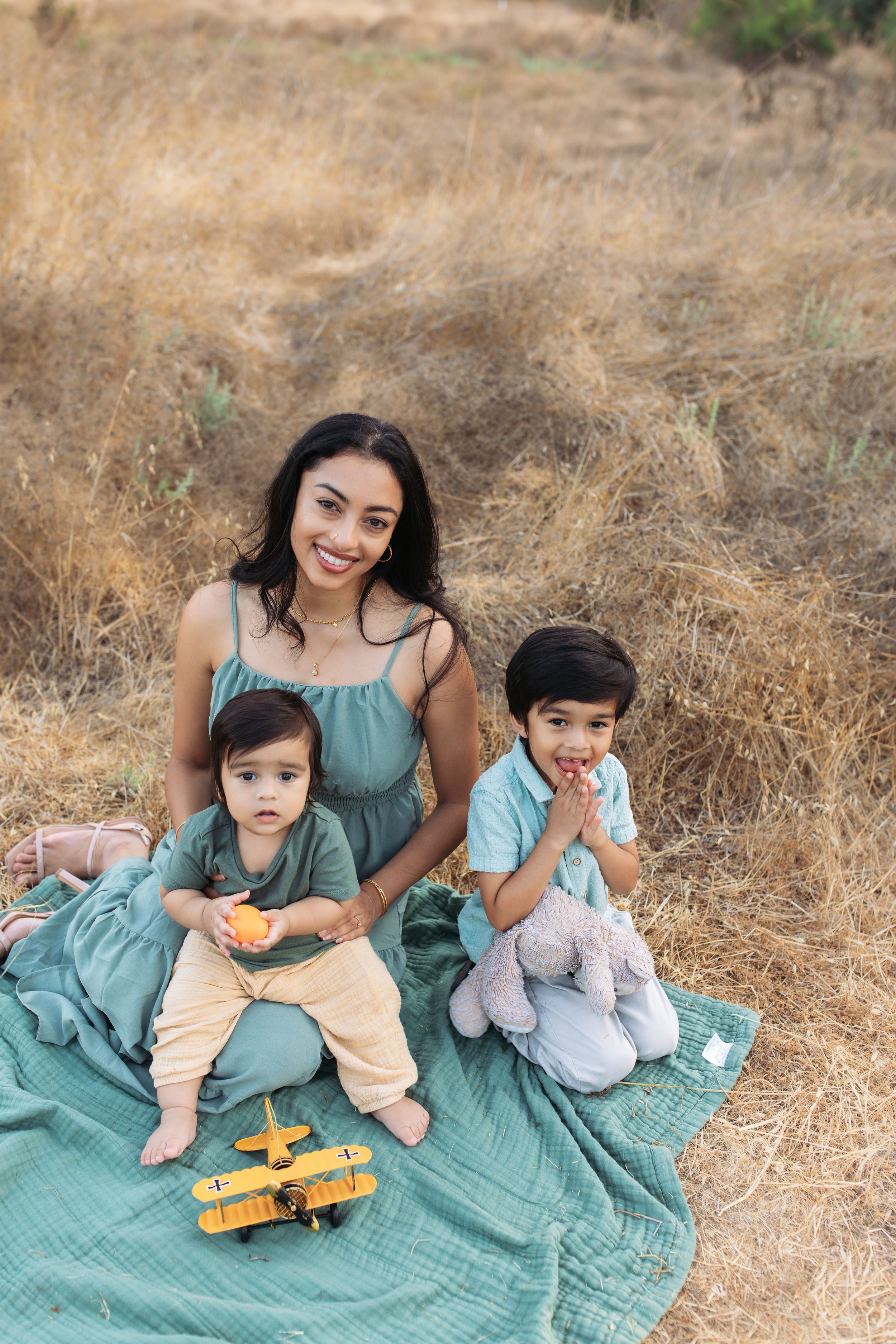 A smiling mother in a green dress sits on a blanket in a dry field with her two young children outdoors.