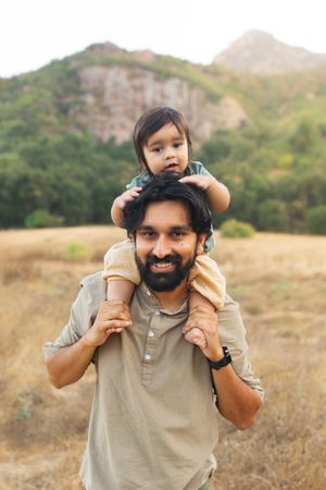 A bearded father smiles while carrying his baby on his shoulders in a scenic outdoor field with mountains.