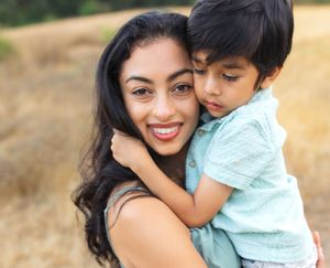 A smiling young woman with long dark hair holds a young boy who hugs her closely in a sunny outdoor field.