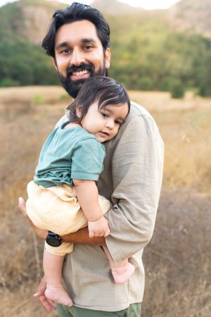 A bearded father smiles while gently holding his baby in a golden outdoor field with mountains in the background.