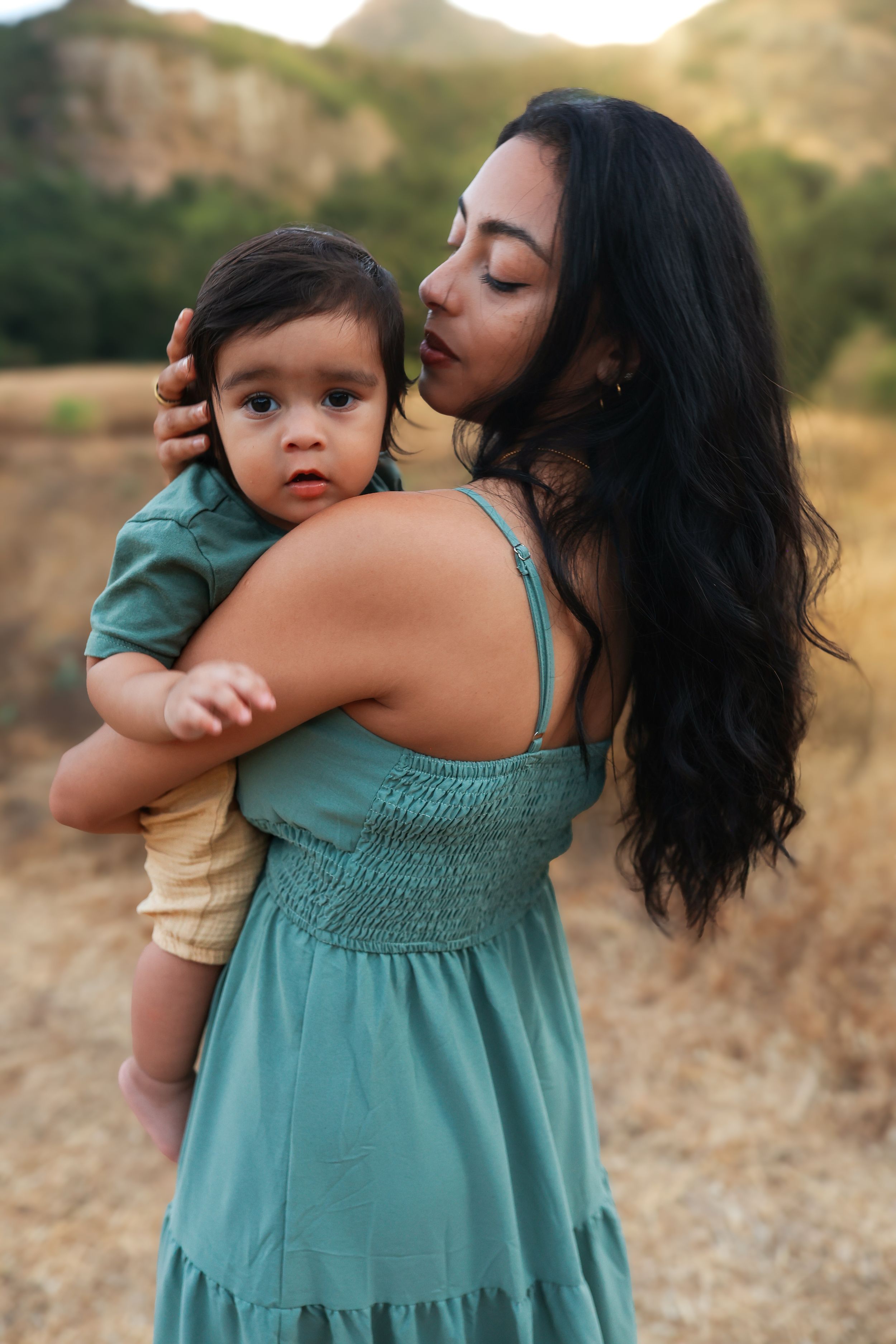 A dark-haired woman in a teal dress tenderly holds and kisses a baby outdoors in a natural field setting.