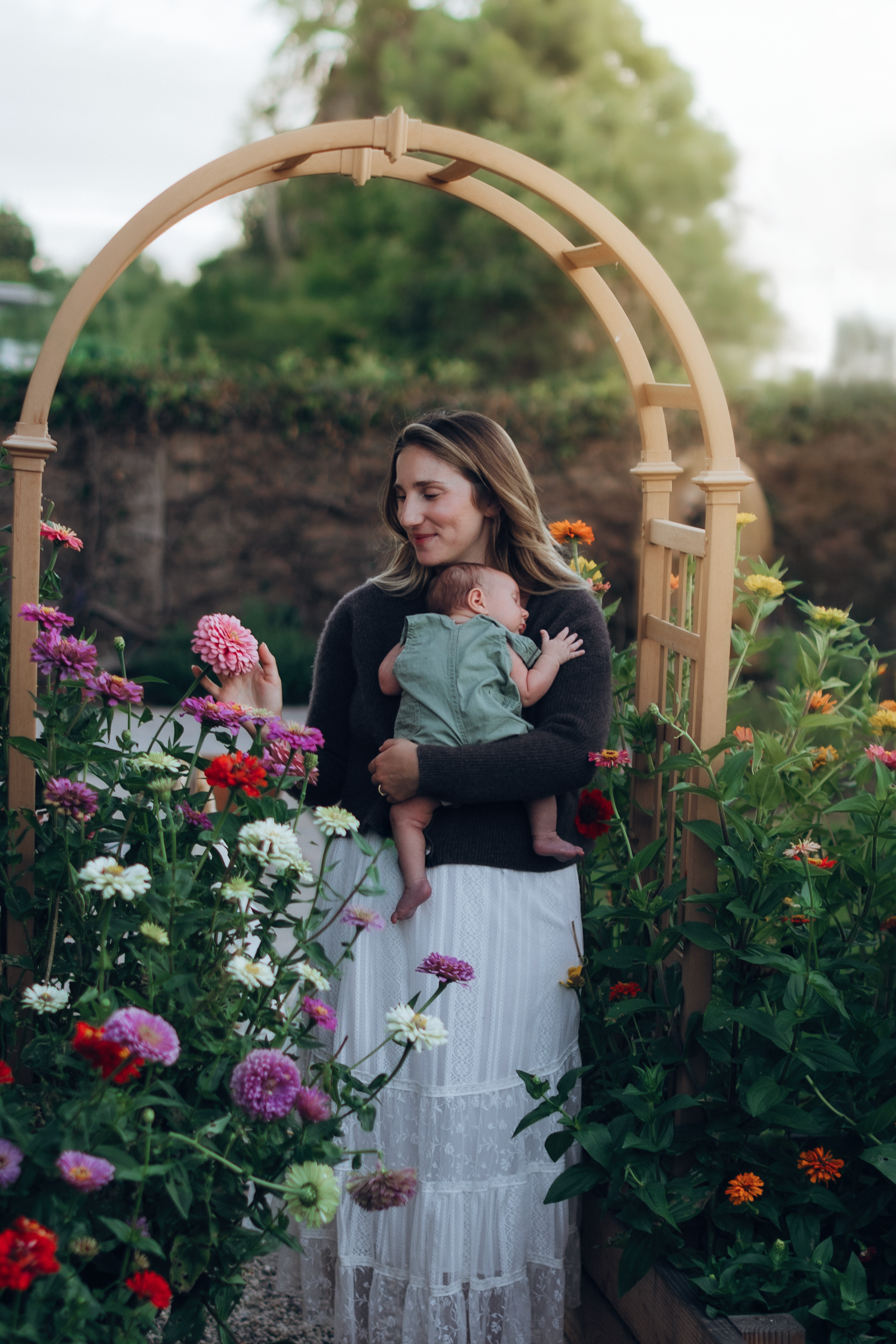 A woman holding a newborn baby stands under a wooden garden arch surrounded by colorful blooming flowers.