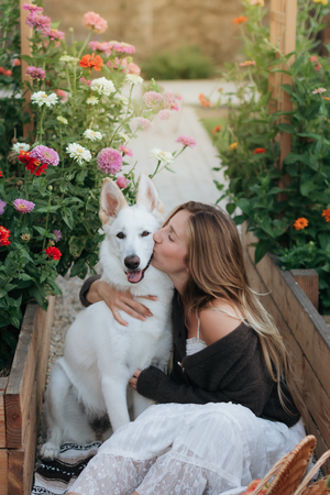 Young woman kissing white Swiss Shepherd dog surrounded by colorful zinnia flowers in a garden.