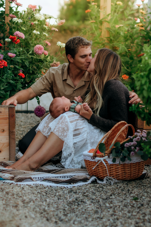 Couple kissing while holding newborn baby during outdoor picnic surrounded by colorful flowers and a wicker basket.