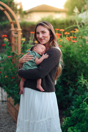 A smiling mother with long hair holds her sleeping newborn baby in a lush garden with orange flowers at golden hour.
