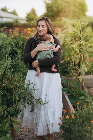 Mother in white skirt and brown sweater tenderly holds newborn baby in lush garden with marigolds.