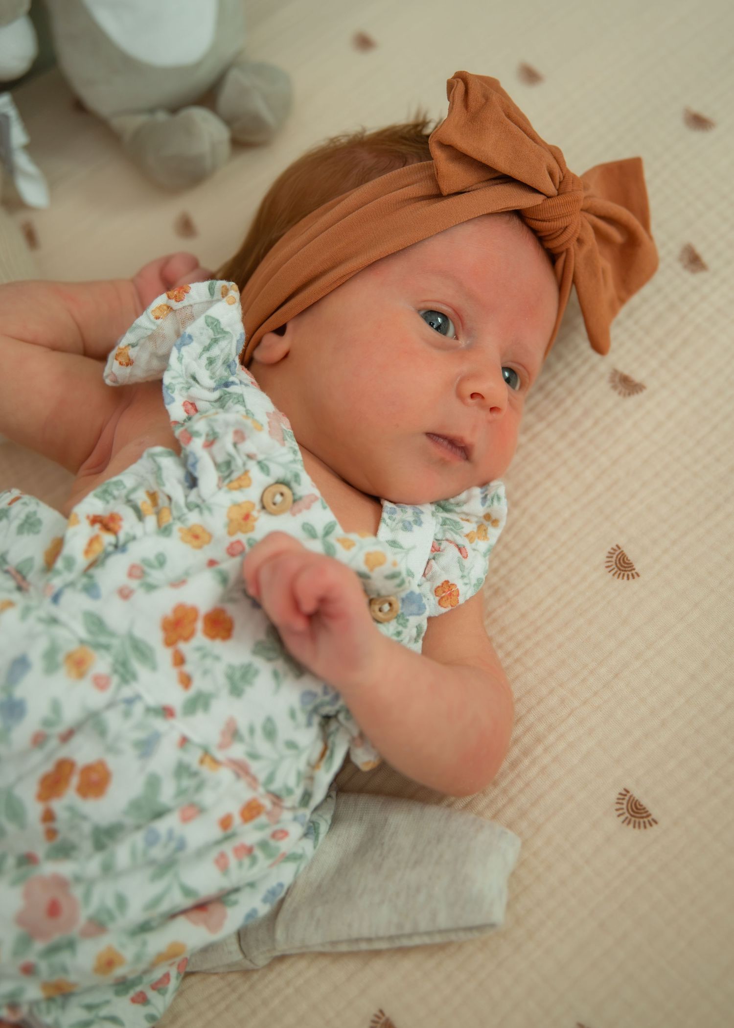 Newborn baby girl in floral romper and brown bow headband lying on a textured cream blanket.