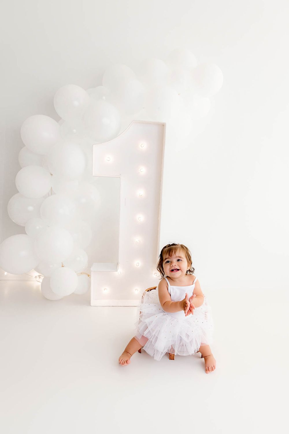 Smiling toddler girl in white tutu dress sits on floor beside illuminated number 1 with white balloon arch for first birthday.