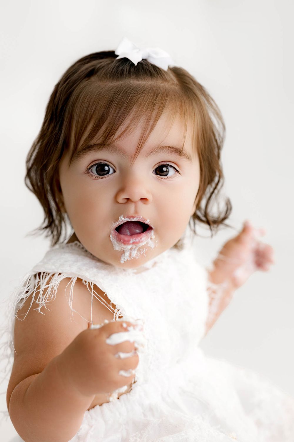 Adorable baby girl in white fluffy dress with frosting on her face during a first birthday cake smash photo session.