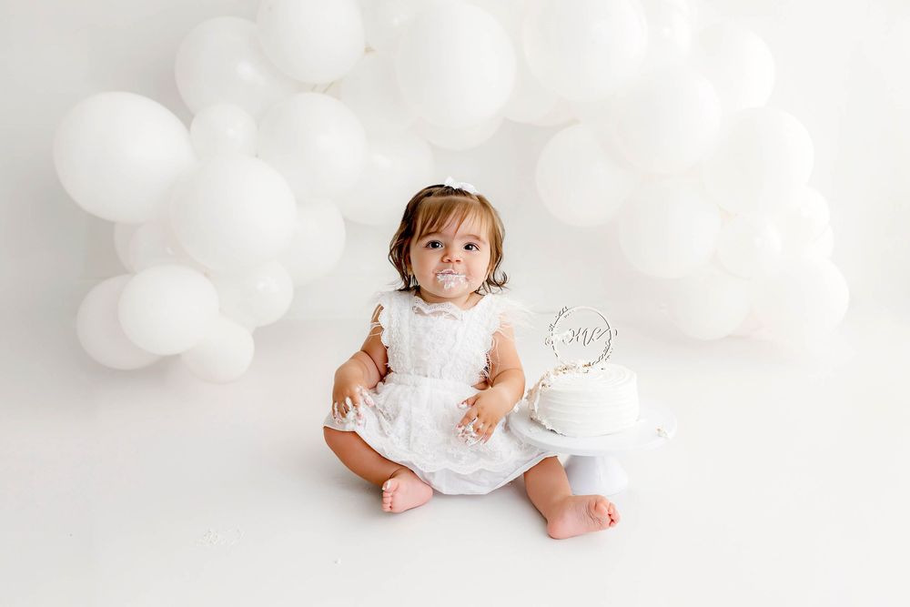 Baby girl in white dress sits by a white cake and balloons during a first birthday photoshoot by French Valley Cake Smash Photographer.