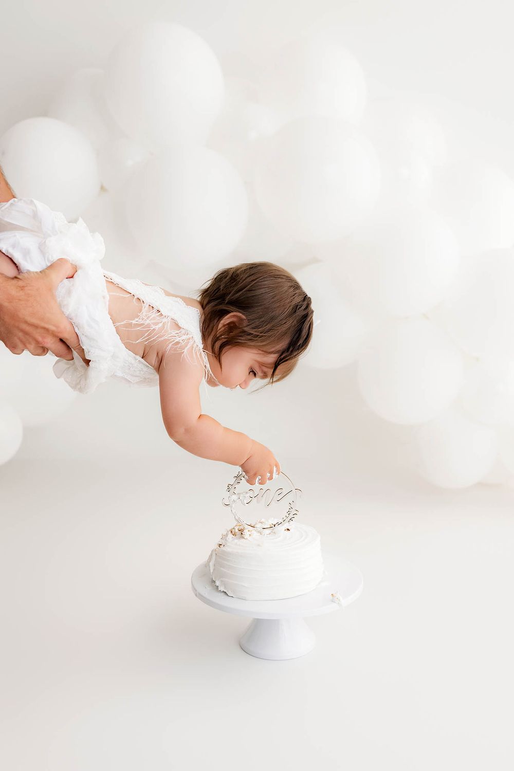 Baby girl reaching for first birthday cake surrounded by white balloons during photo session by French Valley Cake Smash Photographer.