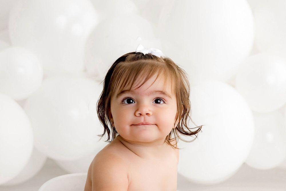 Adorable baby girl with brown hair and white bow headband poses in front of white balloons backdrop.