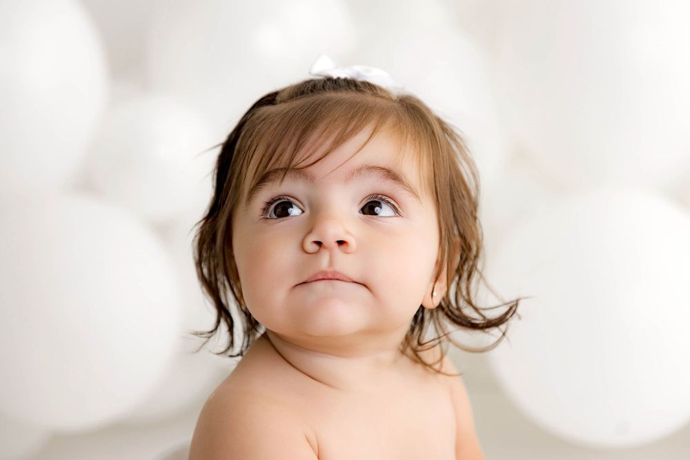 Adorable baby girl with brown hair gazing upward, surrounded by white balloons in a soft, dreamy portrait.
