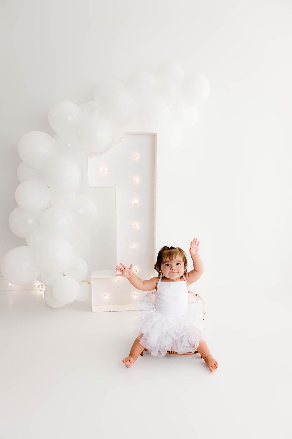 Baby girl in white tutu sits before illuminated number 1 and white balloon arch for first birthday photo.