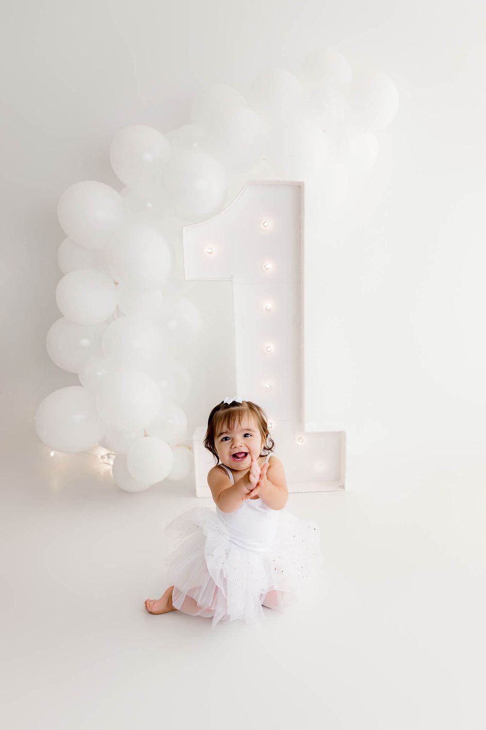 A joyful baby girl in a white tutu sits in front of a illuminated number one and white balloon arch.