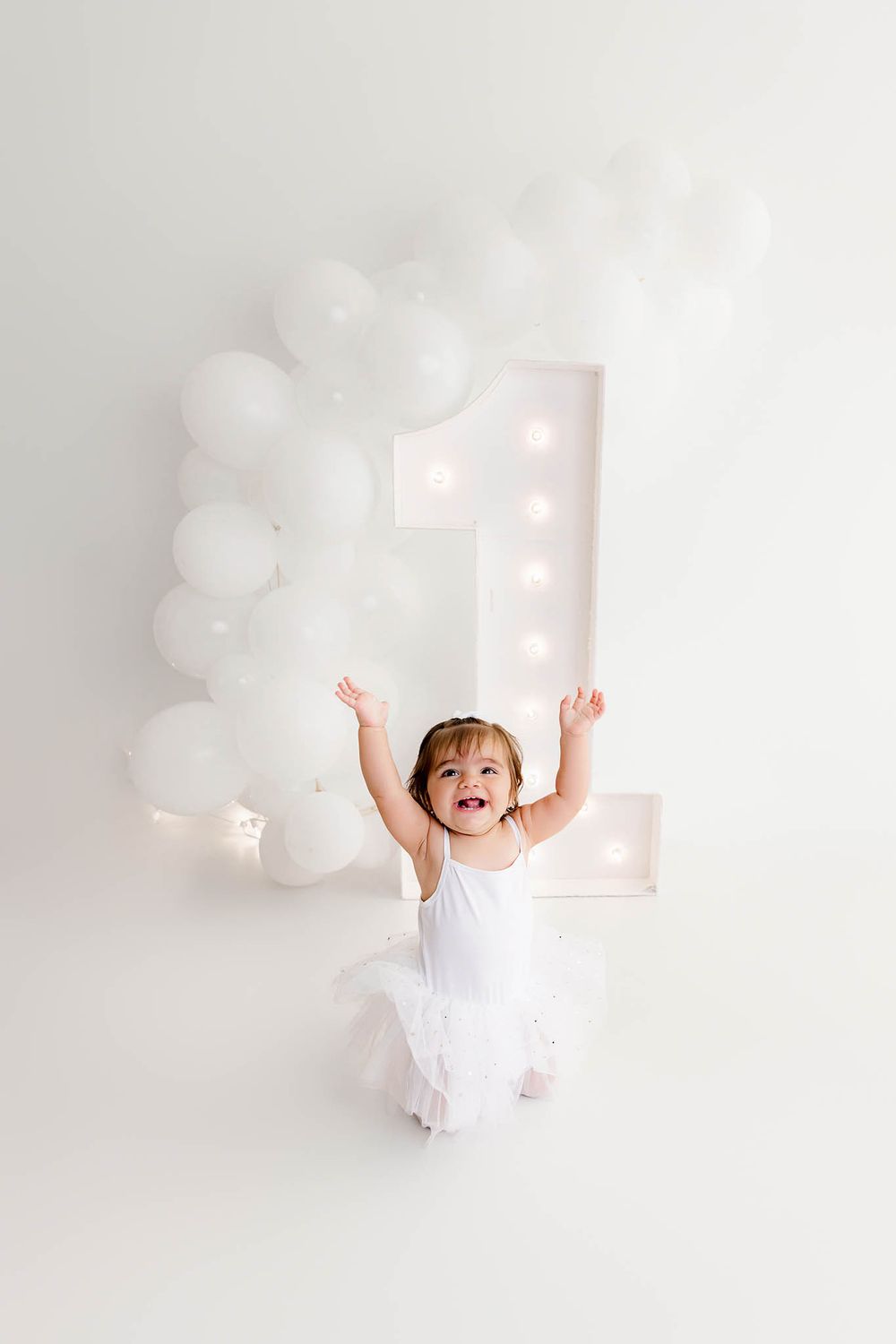 Joyful toddler girl in white tutu celebrates first birthday with arms raised, surrounded by white balloons and marquee number one.
