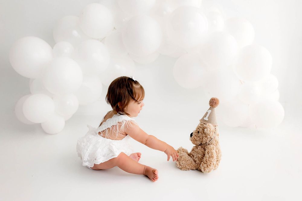 Baby girl in white outfit sits with a teddy bear wearing a party hat, surrounded by white balloons.