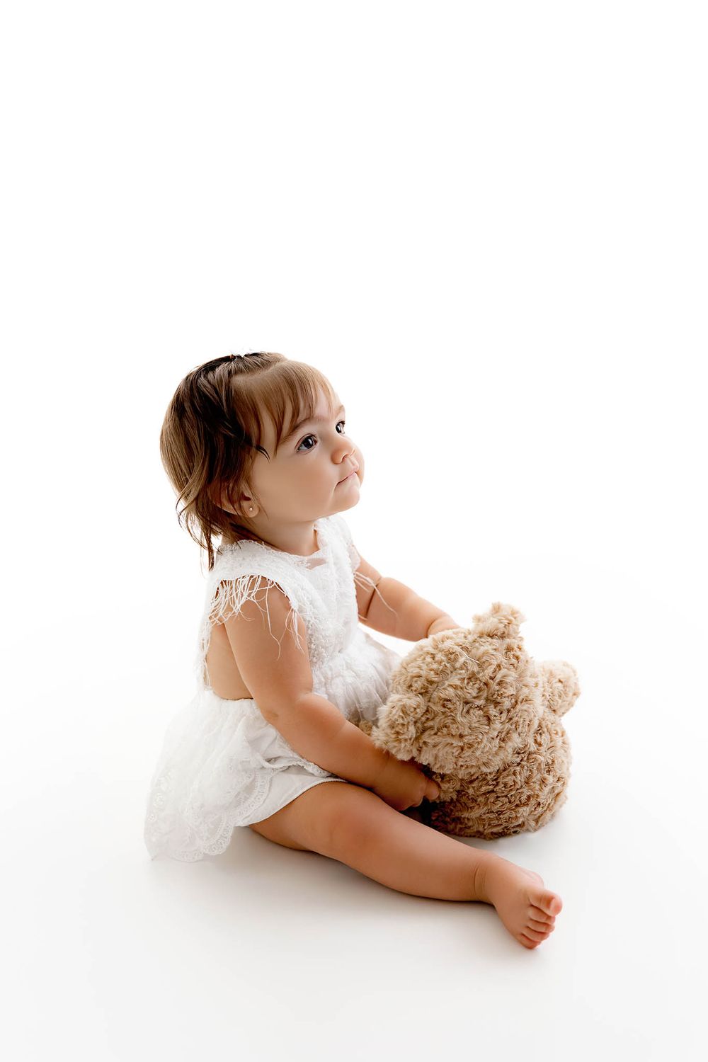 A cute toddler girl in a white lace dress sits barefoot hugging a teddy bear against a white background.