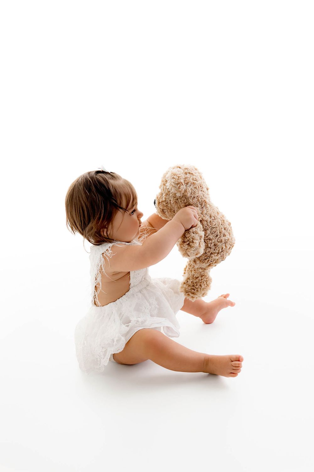 A barefoot toddler girl in a white dress sits on a white floor, holding up a fluffy teddy bear with both hands.
