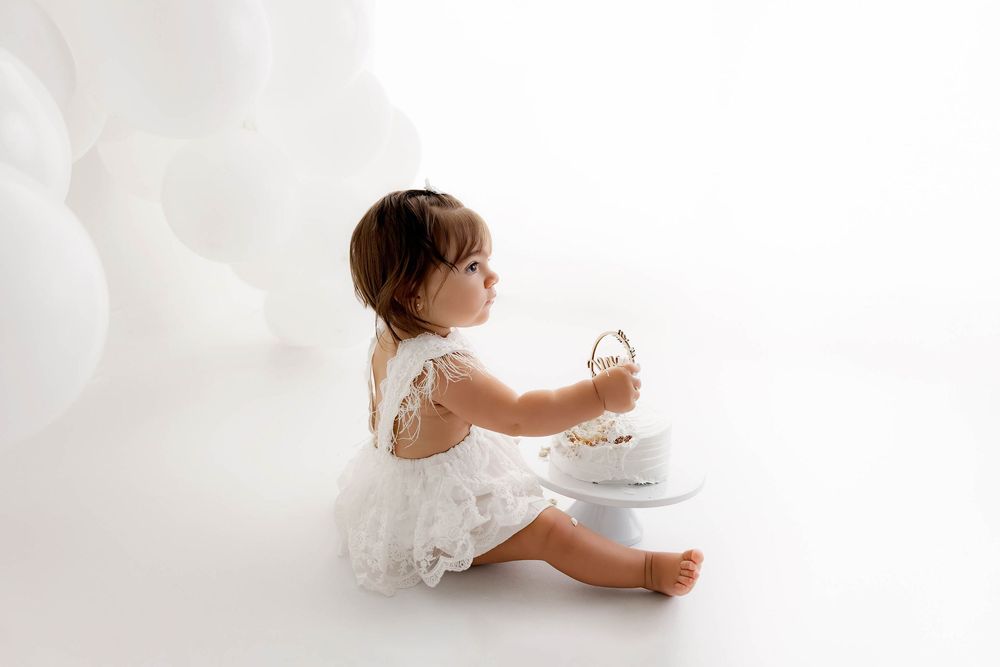 A baby girl in a white dress sits on the floor holding a small cake during a sweet photoshoot by  French Valley Cake Smash Photographer.