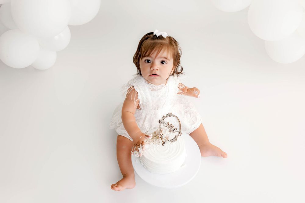Baby girl in white romper sits beside a white first birthday cake, surrounded by white balloon decorations.