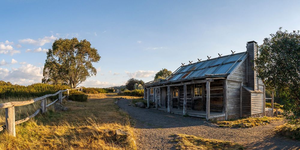 Rustic wooden pioneer hut with corrugated iron roof on a gravel path surrounded by dry grassland and trees under a blue sky.