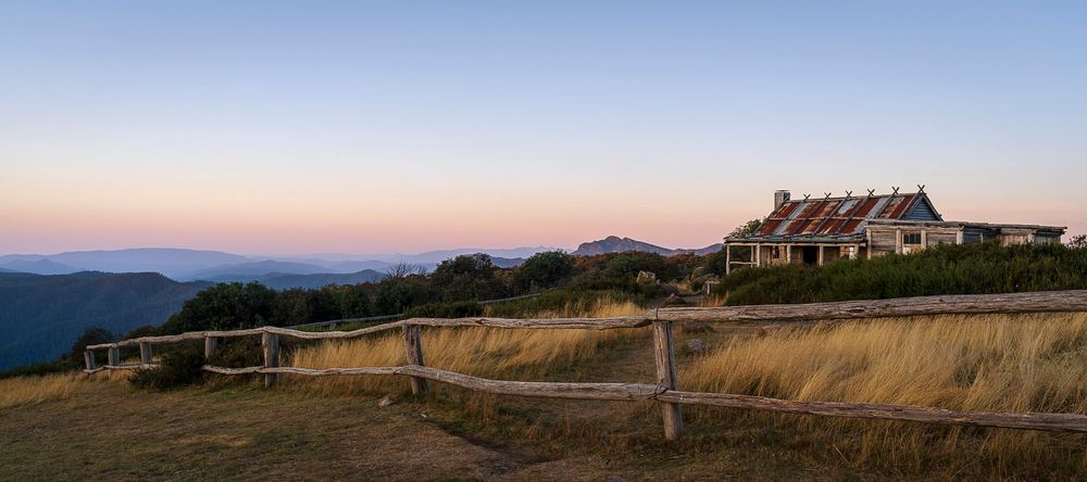 Rustic mountain lodge at dusk with wooden fence, golden grasslands, and blue mountain ranges under a pastel sunset sky.