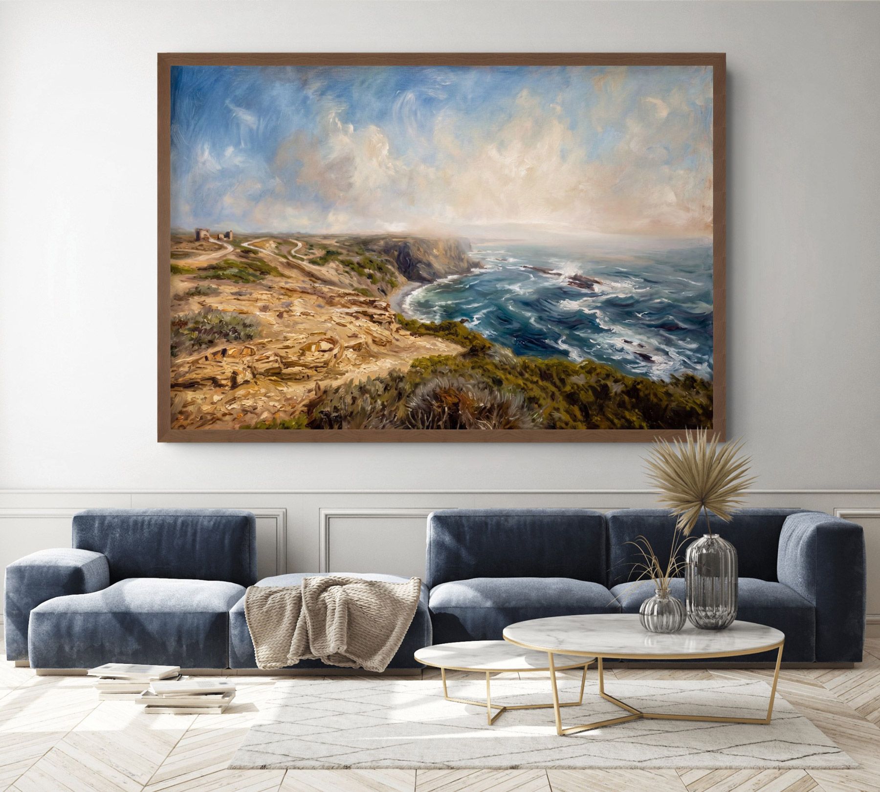 Framed coastal landscape oil painting with blue sofas and gold coffee table in a modern living room.