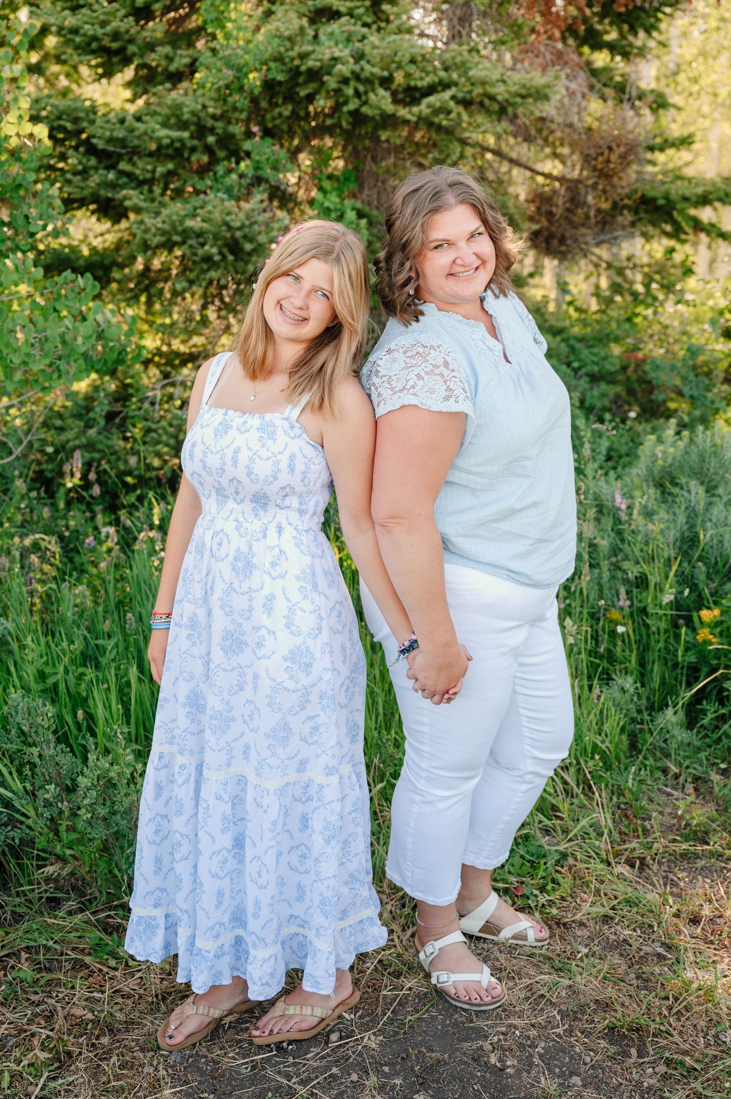 Two women smiling outdoors in white and mint outfits, posing together among lush green trees and foliage.