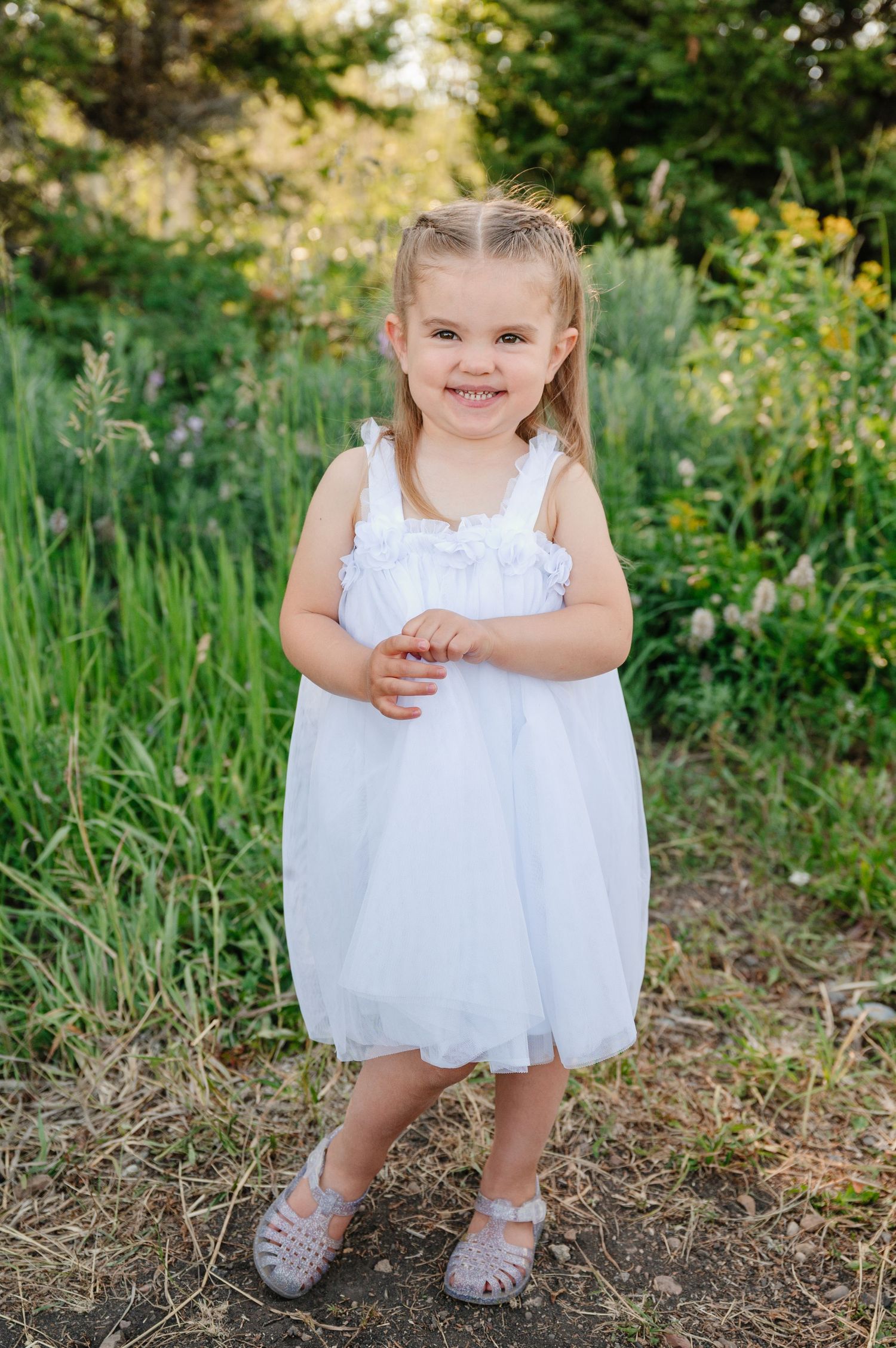 Young girl in white tulle dress and silver sandals smiling outdoors among green plants and wildflowers.
