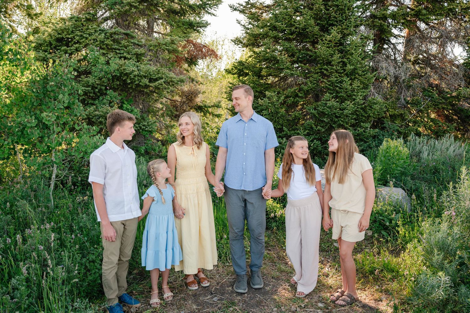 Family of six poses outdoors in a lush garden, wearing coordinated pastel outfits, smiling together on a sunny day.